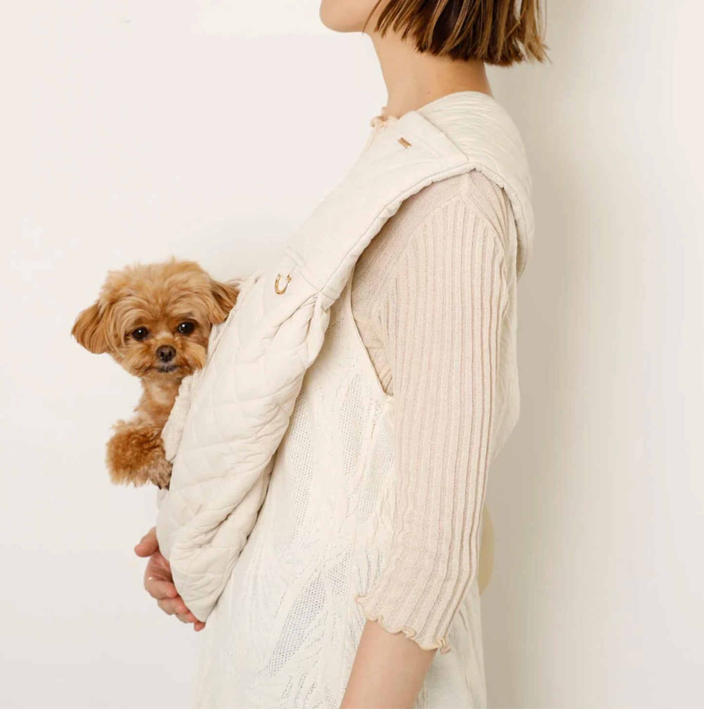 Person wearing a cream-colored knit cardigan holding a small dog on a plain background